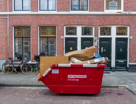 A large red commercial skip with white reflective striping and a warning label is positioned on a paved sidewalk in front of a row of three brick terraced houses. The skip is filled with mixed waste materials, including large flattened cardboard boxes, plastic bags, and various packaging debris, all stacked haphazardly. To the left of the skip, two bicycles are parked and secured to a bike rack, with one leaning against the others. The background features a typical urban residential street scene with brick facades, ground-floor windows with metal grilles, and front doors painted dark green, all under a clear sky. The presence of the skip and waste suggests a rubbish removal service, possibly handled by Commercial Waste Oxford, servicing local homes or small businesses in the area near Oxford. The scene captures an everyday waste collection environment in a residential town setting, emphasizing proper management of rubbish disposal on a typical street.