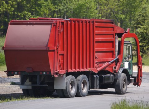 A large red commercial waste disposal truck with a sturdy metal container parked on a paved surface, surrounded by greenery with trees in the background. The container has vertical ridges and a smooth, angled front panel designed for collecting and transporting rubbish. The truck features a cabin with a side door and a side mirror, with black rubber tires on silver rims. The environment appears bright and clear, indicating daytime, with the truck positioned in an outdoor setting that could be typical for rubbish collection services around Oxford or nearby areas. This image exemplifies professional waste removal equipment used by Commercial Waste Oxford for rubbish collection and disposal services in the local region.