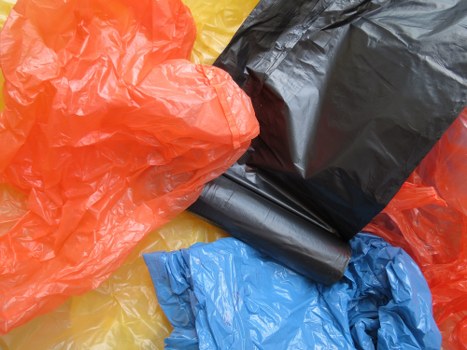 A close-up view of several tightly crumpled plastic rubbish bags in various colours, including bright orange, black, blue, and yellow, piled together on a surface. The bags appear to be made of thin, flexible plastic material with a glossy finish, and their surfaces show typical crinkled textures. The black bag is prominent and partially piled on top of the others, with the orange and blue bags lying nearby. The setting suggests an outdoor area or driveway, possibly related to waste collection or rubbish removal services in Oxford, reflecting typical materials handled by Commercial Waste Oxford. The background is not visible, focusing solely on the different coloured plastic bags gathered together, indicating they are ready for disposal or collection.