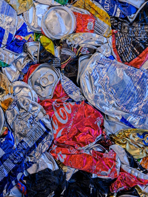 A close-up view of a large pile of crushed and crumpled aluminium cans and plastic bottles, primarily in silver, red, blue, and yellow colors. The cans display visible pull tabs, textured surfaces wit