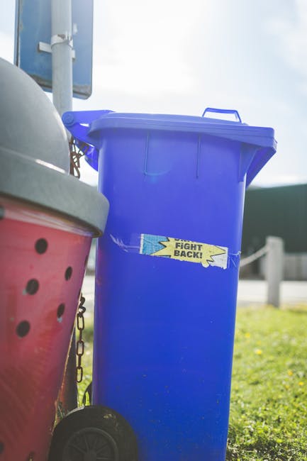 A close-up view of a large pile of crushed and crumpled aluminium cans and plastic bottles, primarily in silver, red, blue, and yellow colors. The cans display visible pull tabs, textured surfaces wit